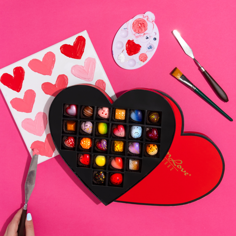 24 beautifully crafted chocolates in a red, heart-shaped gift box. The gift box includes 10 limited -edition hearts as well as 14 signature pieces. The box is displayed on a pink background with a paintbrush and pink and red painted hearts.